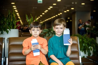 Two Young Kids Sitting On Brown Chairs Holding Passports And Tickets In Waiting Hall In Airport